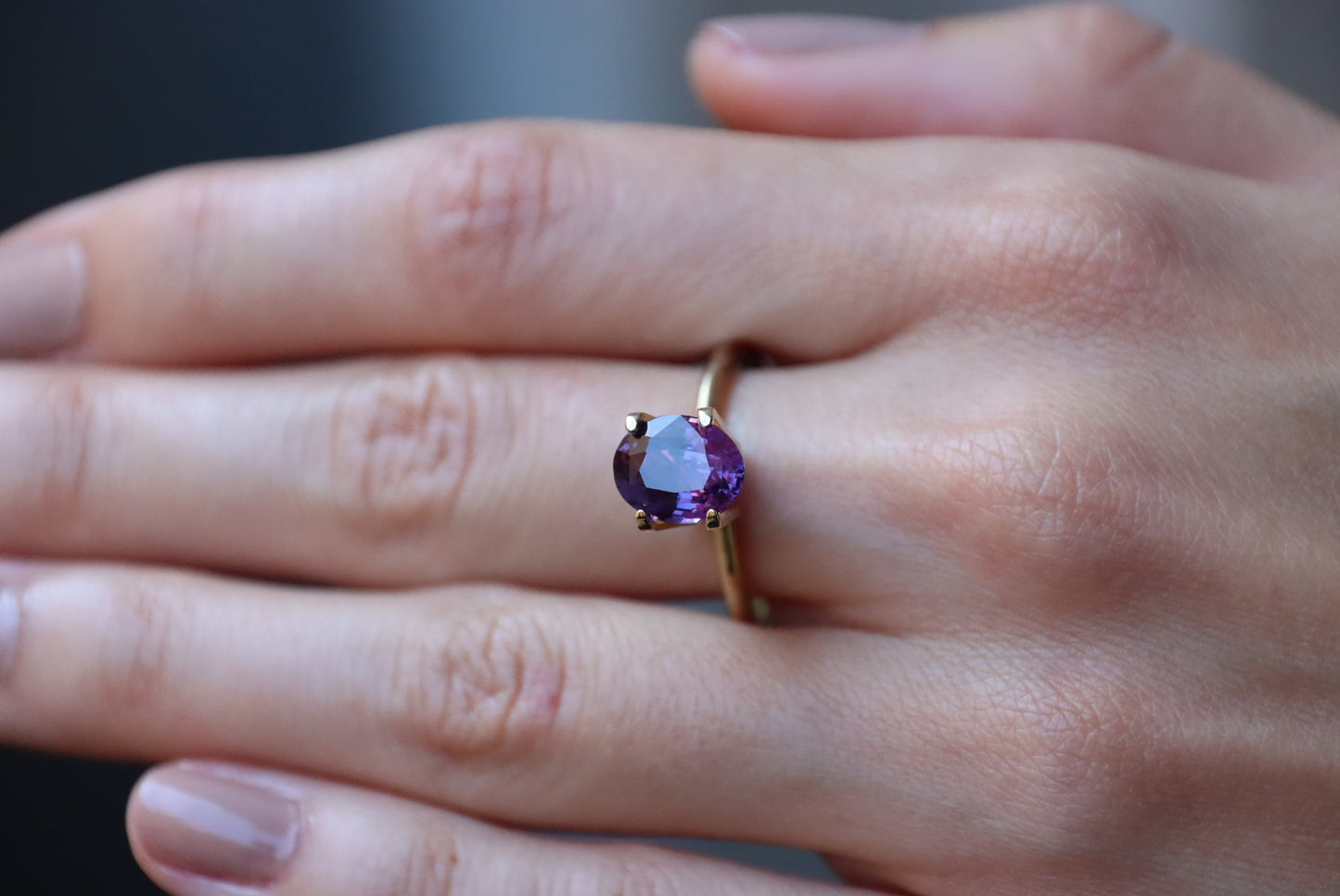 Une main au vernis à ongles nude revêt la bague en or Bonnot Paris « Saphir Violet · 2,61 ct », mettant en valeur une pierre précieuse violette taille ovale sur l'annulaire, sur un fond flou.