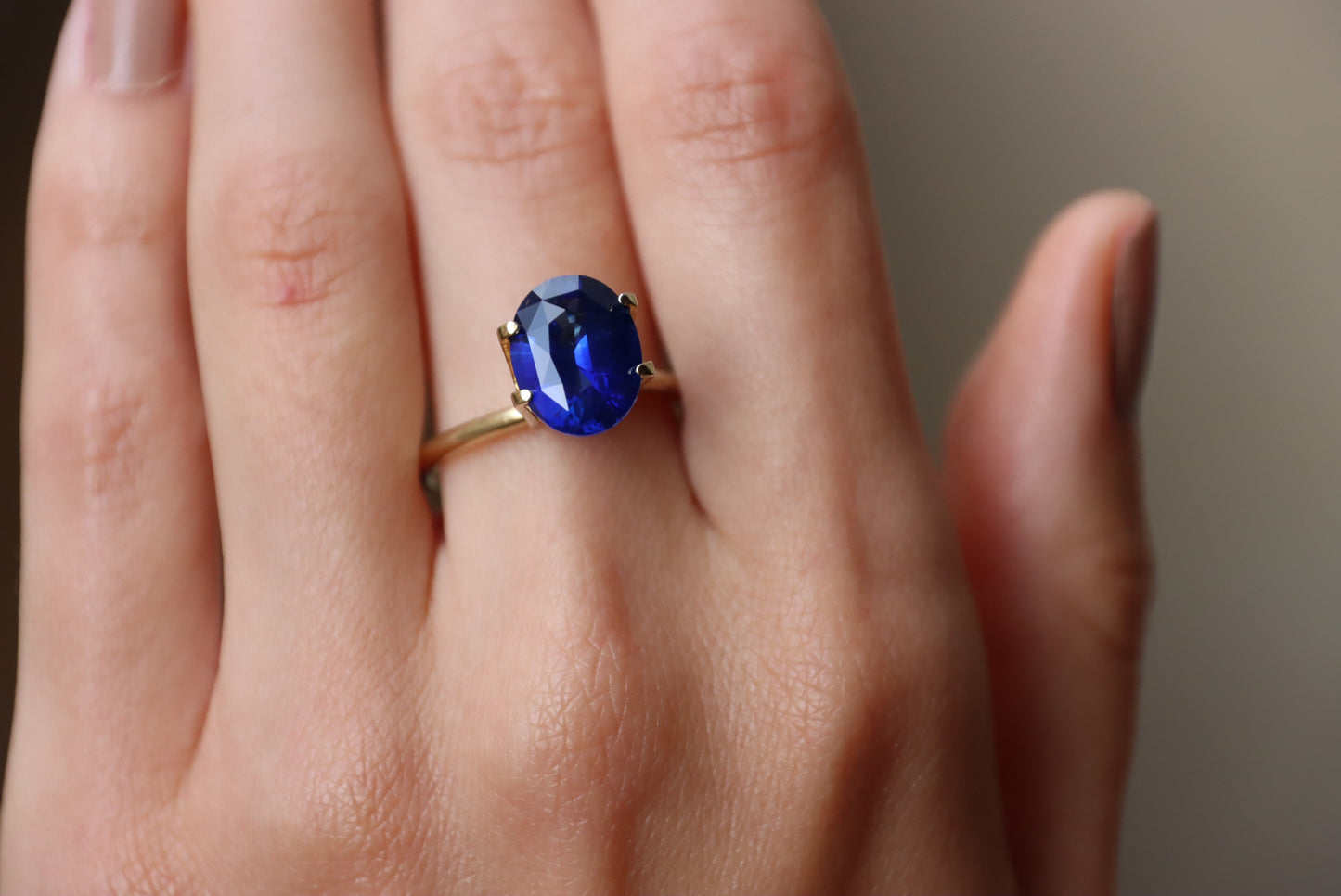 Une main ornée de la bague Saphir Bleu · 4,05 carats de Bonnot Paris, ornée d'une pierre ovale bleu profond sertie en son centre, est présentée sur un fond neutre. Les doigts paraissent détendus, les ongles soigneusement taillés.