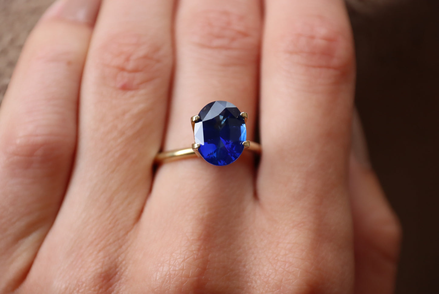 Gros plan d'une main ornée du Bonnot Paris "Saphir Bleu · 4,05ct", comportant une bague en or avec une grosse pierre précieuse bleue ovale sur l'annulaire sur un fond flou.