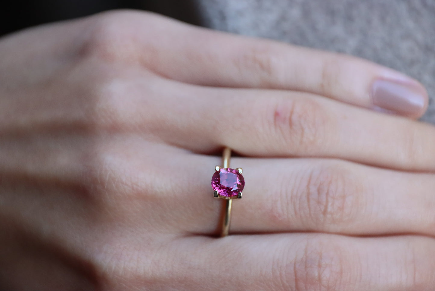 Une main ornée d'une bague en or 1,68 carat Rubis de Birmanie de Bonnot Paris, ornée d'une pierre rose ovale, repose délicatement sur un fond de tissu gris. Les ongles sont soigneusement manucurés avec un vernis naturel clair.
