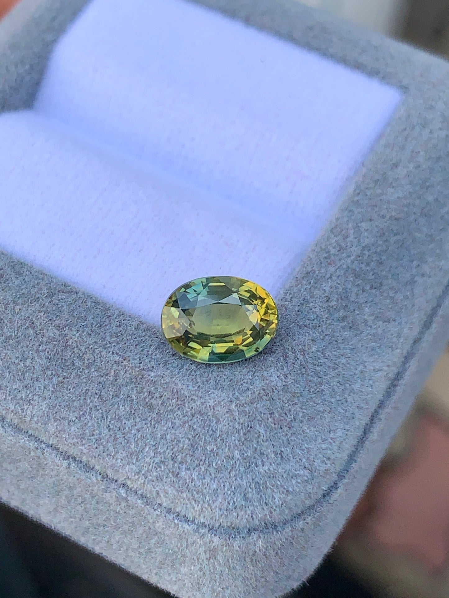Le Saphir Antique · 2,56 ct de Bonnot Paris est une pierre précieuse bicolore taille ovale étincelante, affichant des teintes vertes et jaunes avec une brillance impeccable, sertie dans une boîte à bijoux grise ouverte doublée de tissu blanc.