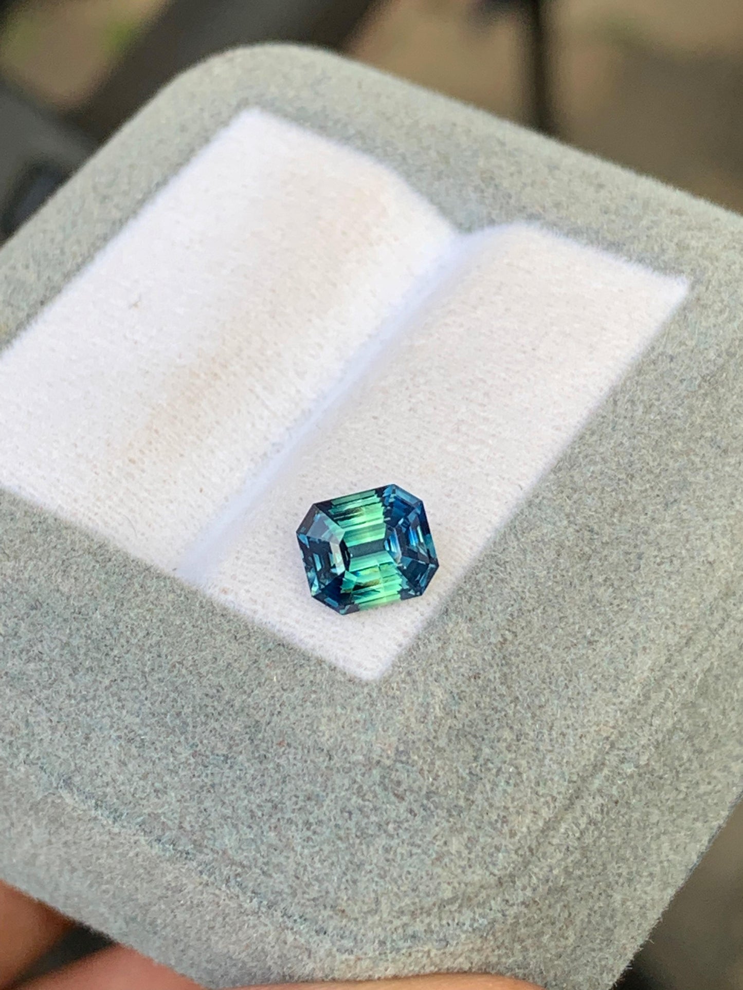 Le Saphir Teal de Bonnot Paris est une pierre précieuse taille émeraude scintillante de 1,65 carat. Logée dans un écrin doublé de velours gris, ses facettes révèlent des teintes captivantes allant du bleu profond au vert.