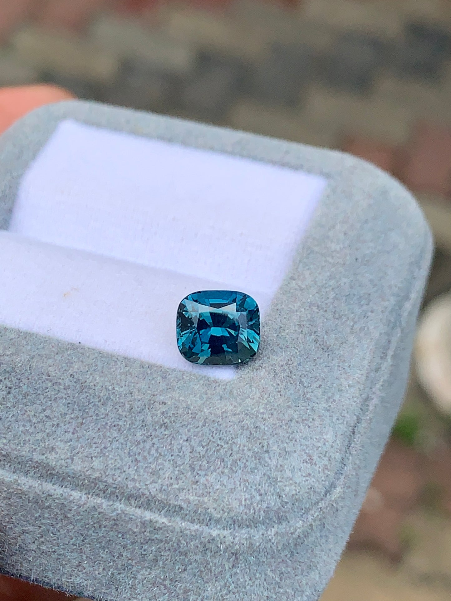 Le Saphir Teal • 2,50 carats de Bonnot Paris, un saphir taille coussin de 2,50 carats, est élégamment présenté dans un écrin de velours gris clair. Un fond légèrement flou met en valeur les détails exquis de ce trésor de Madagascar sur une surface aux motifs subtils.