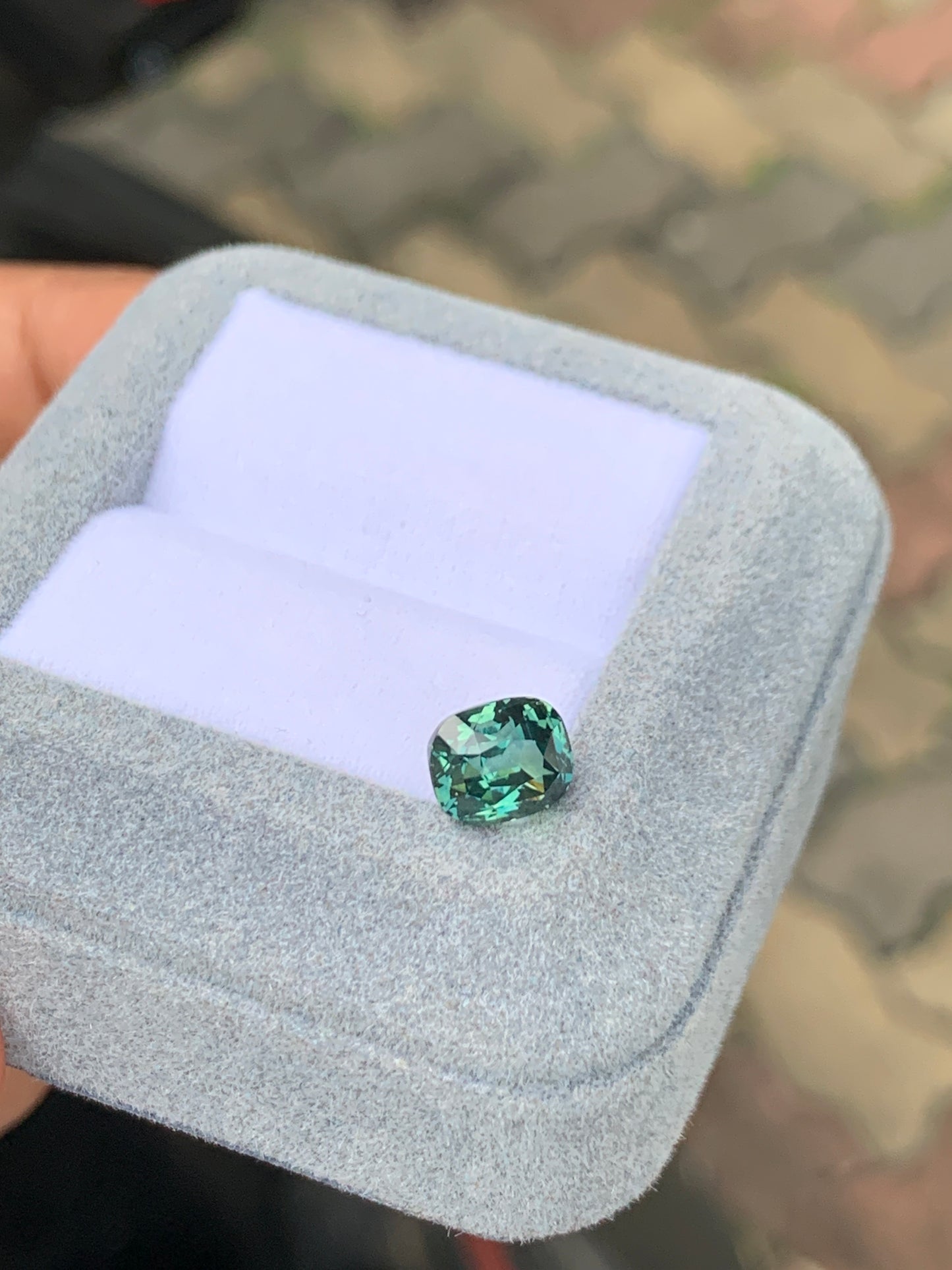 Un Saphir Teal taille coussin de 2,53 ct, de Bonnot Paris, est présenté dans un coffret à bijoux en velours gris clair avec un intérieur blanc sur un fond de trottoir flou.
