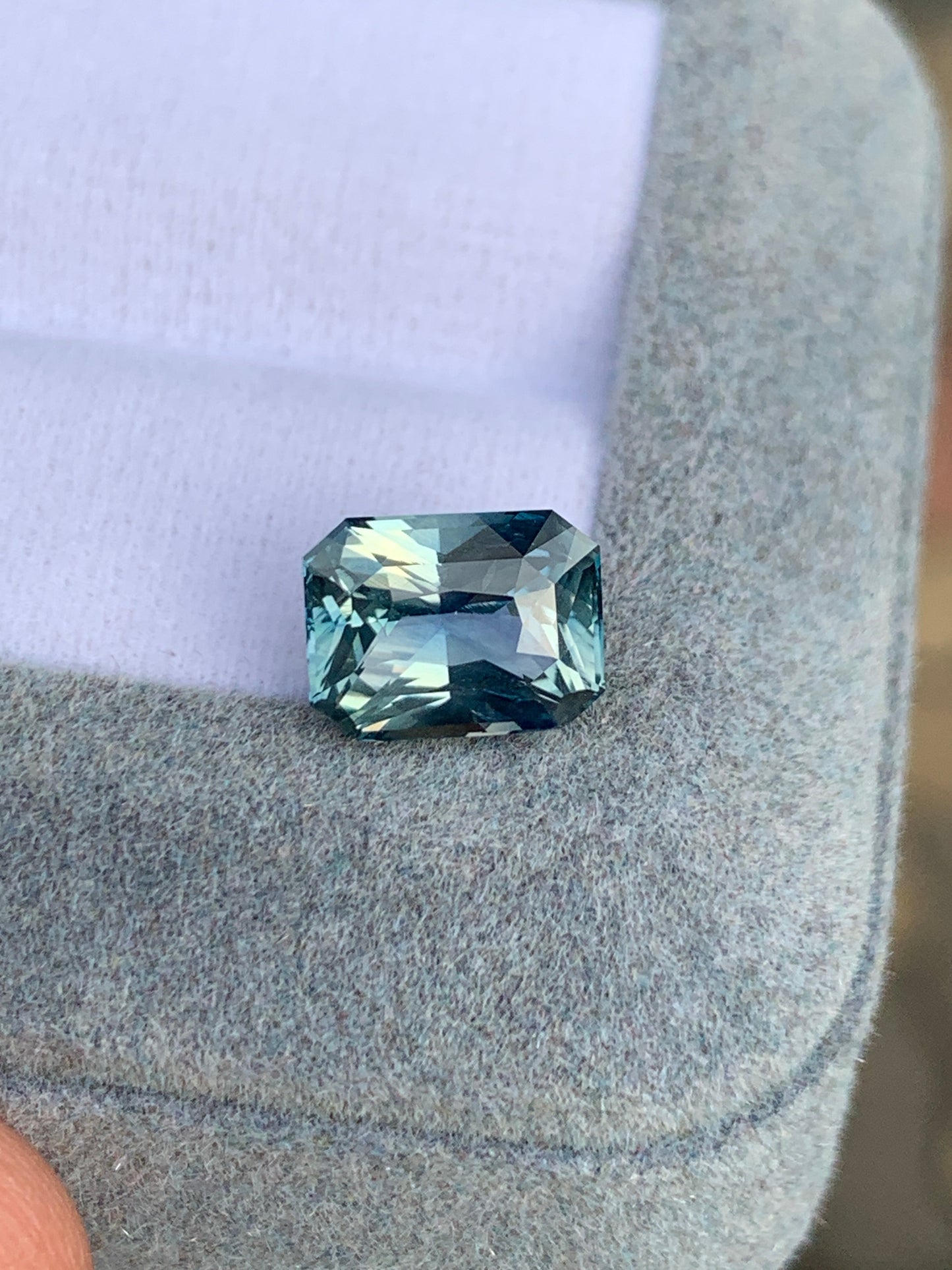 Une pierre précieuse Saphir Teal de 3,20 carats de Bonnot Paris, originaire de Madagascar, repose sur une surface gris clair et douce. Ses facettes rectangulaires scintillent, captant la lumière pour révéler de magnifiques teintes variées.