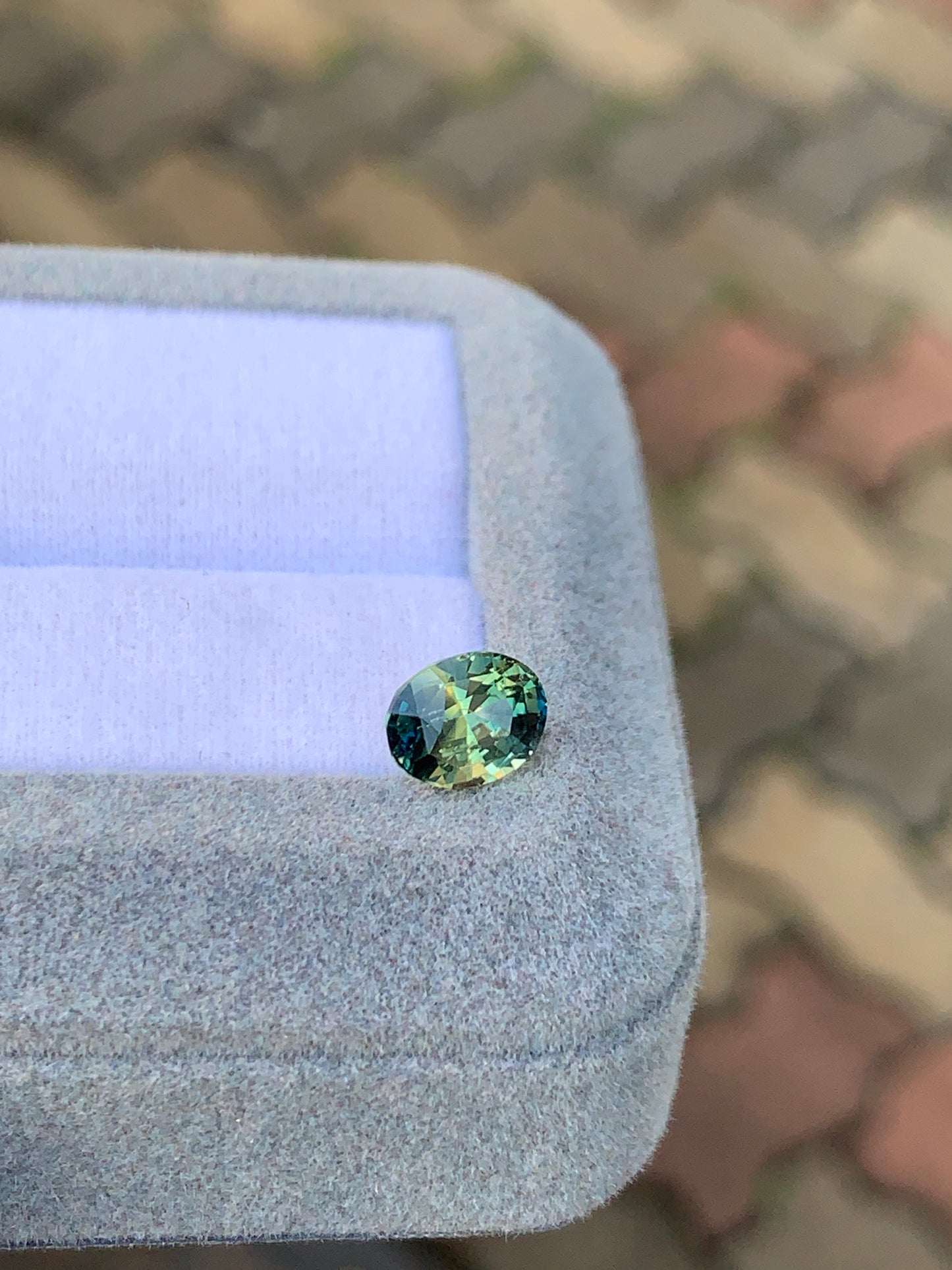 Un saphir ovale bicolore de 2 carats de Bonnot Paris, aux reflets verts et bleus, repose sur un écrin en velours gris clair. Le fond est un motif brique flou aux tons neutres, symbole d'une joaillerie raffinée.