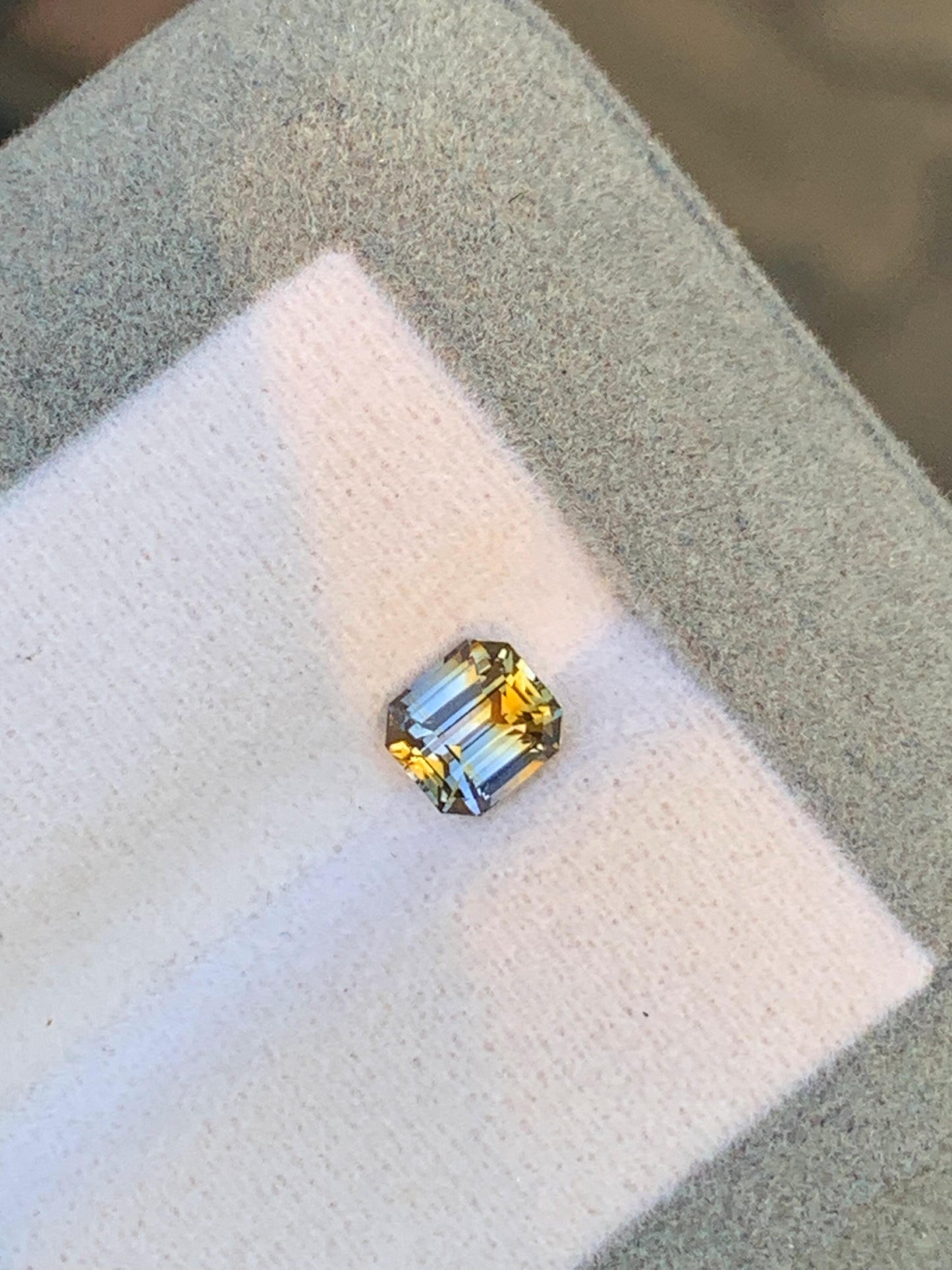Un saphir bicolore de 1,03 carat de Bonnot Paris, taille émeraude carrée, présente des reflets bleus à jaunes et repose sur une surface gris clair. Ses facettes reflètent la lumière, mettant en valeur ses couleurs vibrantes et sa clarté – une véritable merveille parmi les pierres précieuses naturelles.