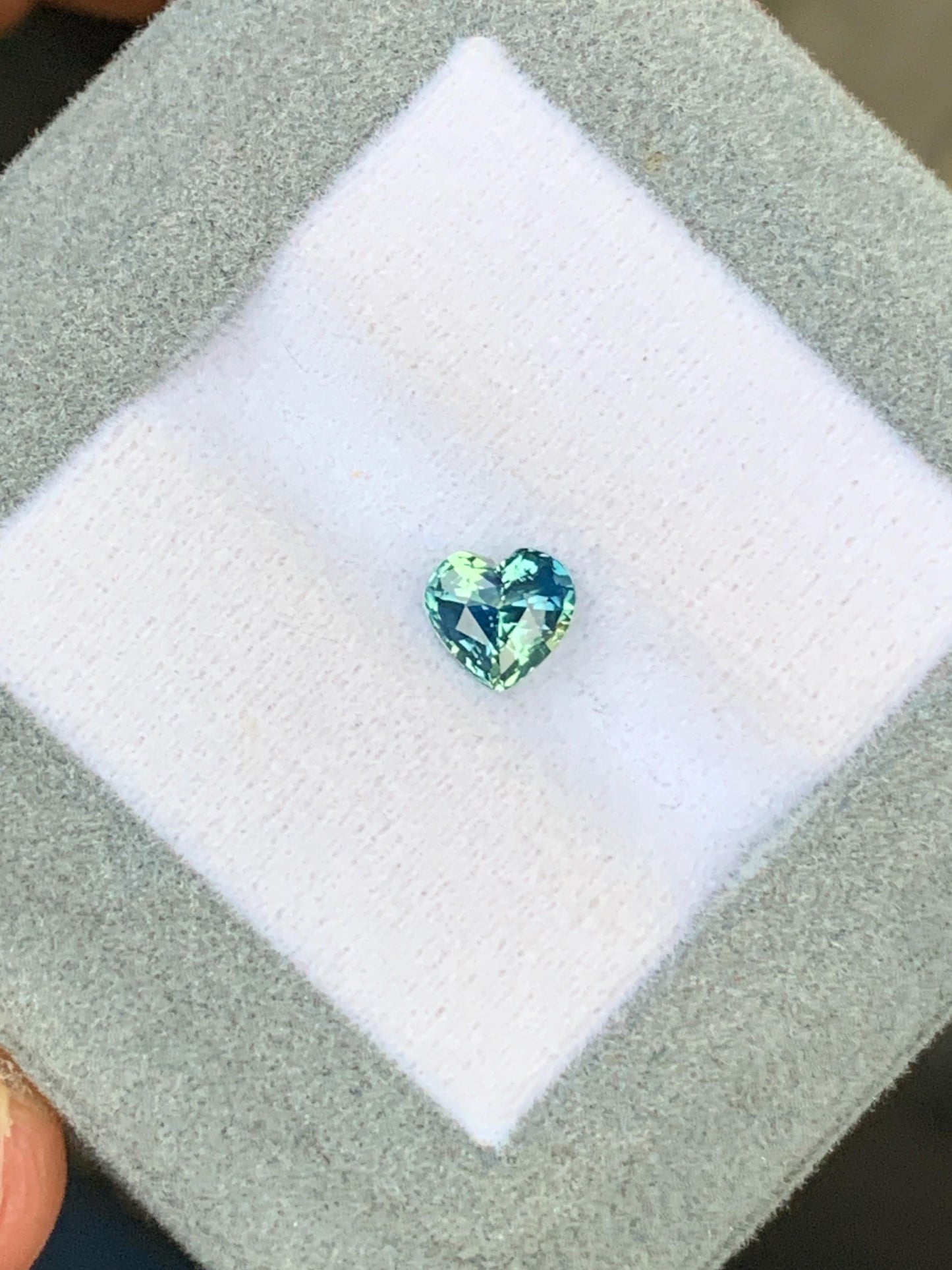 Un saphir bicolore en forme de cœur de 1,06 ct bleu et vert de Bonnot Paris, niché sur un coussin blanc à l'intérieur d'une boîte grise, provient du Sri Lanka.
