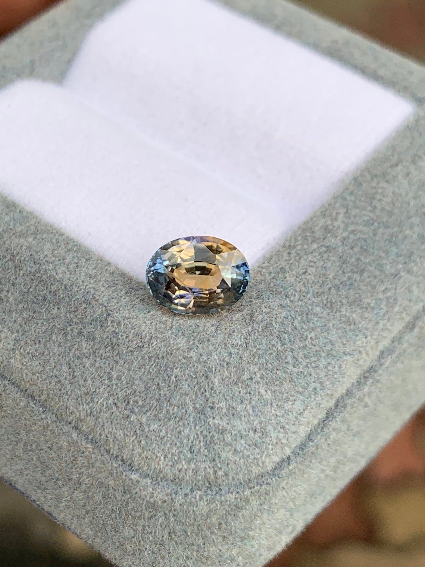 Un Saphir bicolore • 2,02ct de Bonnot Paris, aux teintes ovales de bleu, vert et marron clair, repose sur une surface grise texturée avec un fond légèrement flou.