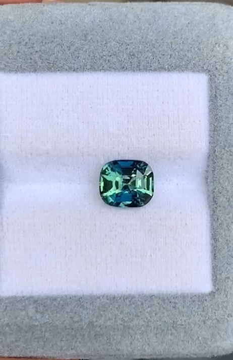 Une pierre précieuse bleu-vert de forme carrée de Bonnot Paris, semblable à un Saphir bicolore • 2,50ct, repose sur un coussin blanc avec un fond de bordure gris doux.