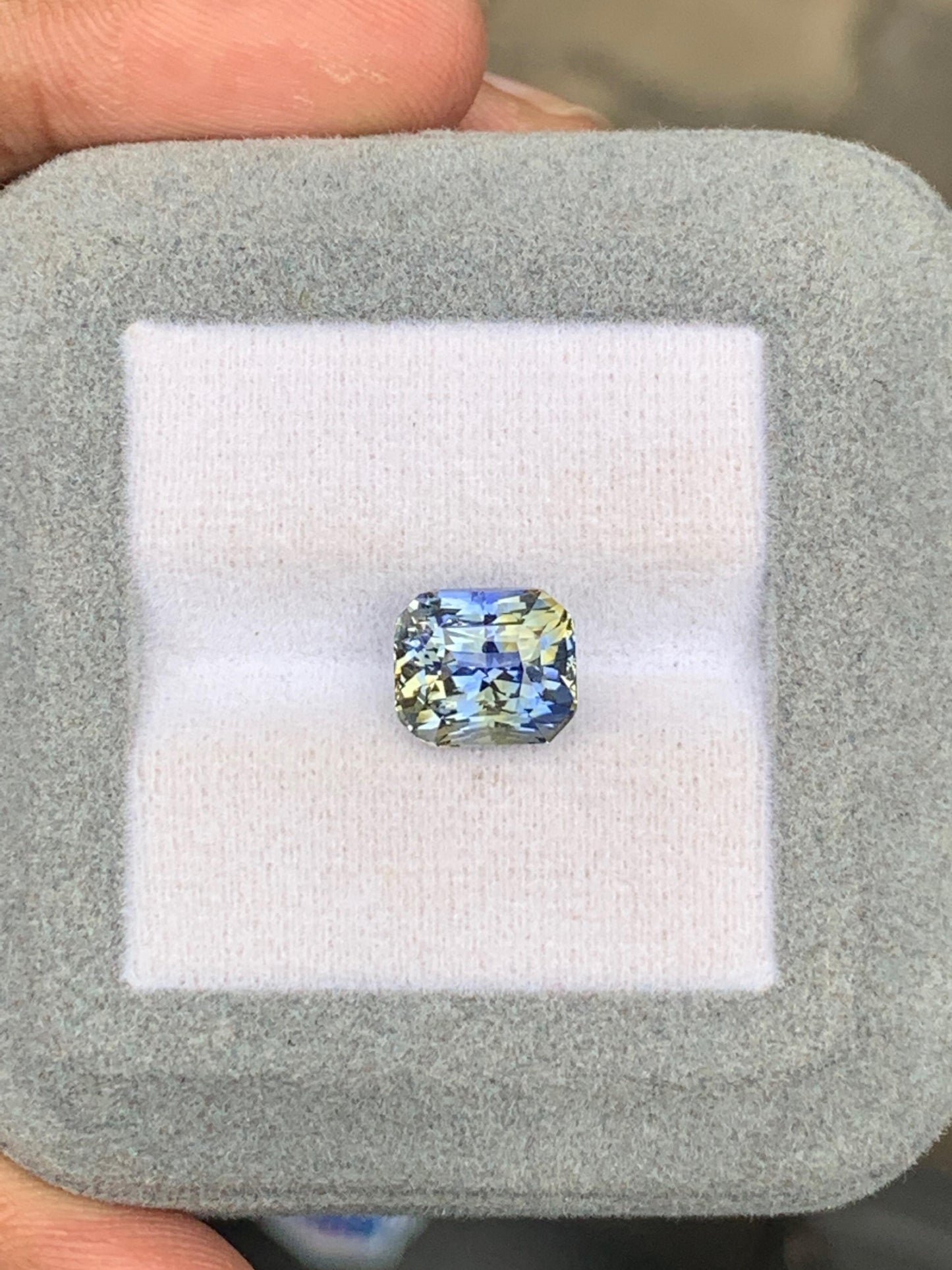 Un saphir bicolore de 5,01 carats signé Bonnot Paris, aux reflets bleu-jaune, est niché dans un écrin gris doux. Des doigts délicatement saisis mettent en valeur les facettes complexes de la pierre, évoquant les éclatantes pierres précieuses du Sri Lanka.