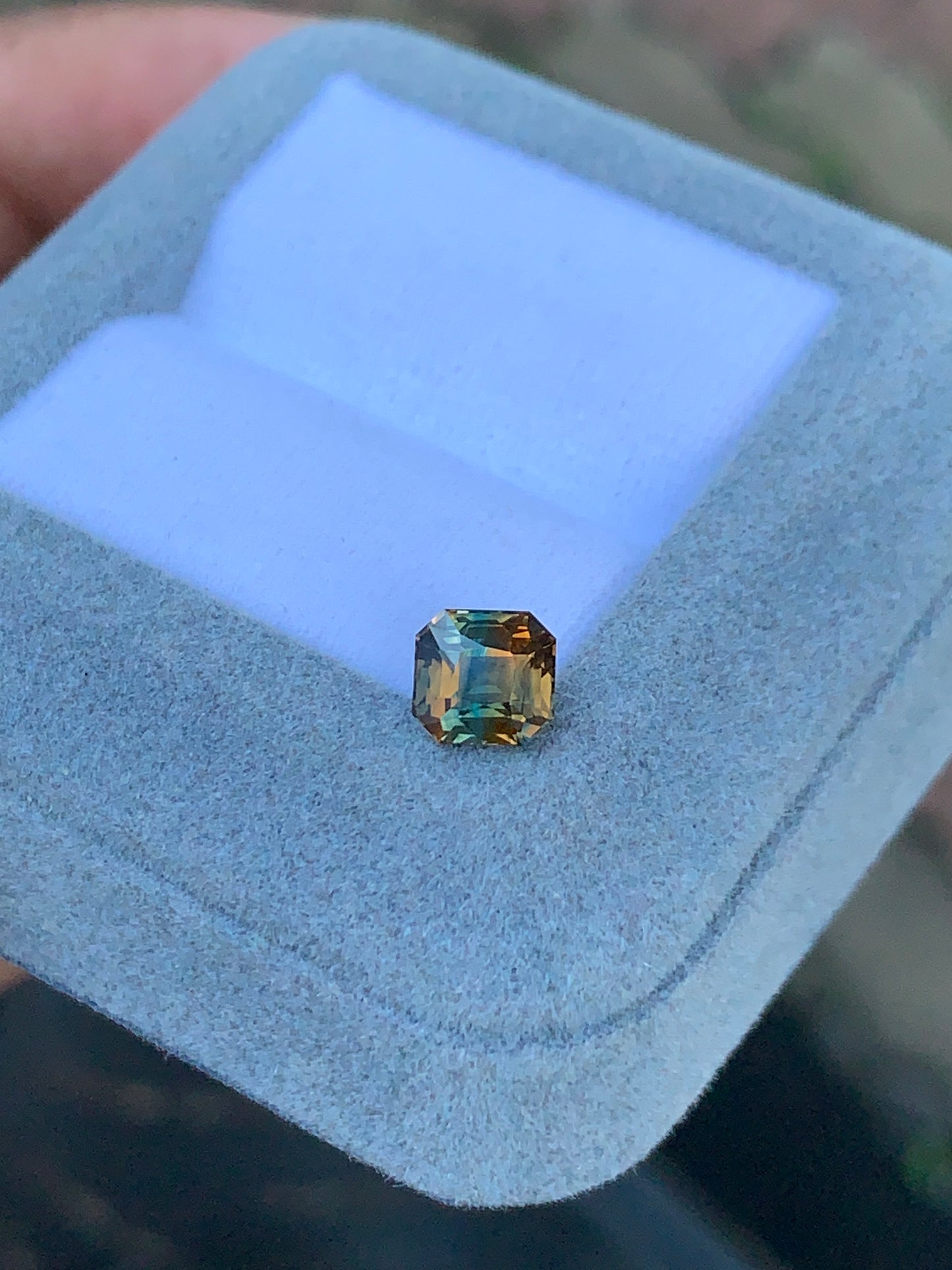 Une main tient un luxueux coffret à bijoux en velours bleu pâle signé Bonnot Paris, exposant le Saphir bicolore de 1,60 carat. Cette pierre taille émeraude présente des nuances vertes et brunes ; ses facettes captent la lumière, révélant une clarté et une vivacité rappelant une forêt d'un vert intense teinté d'orange chaleureux.