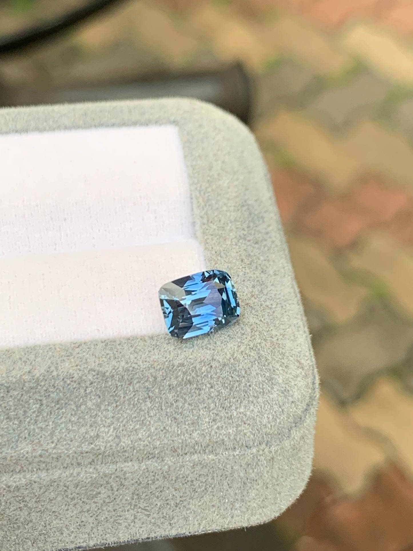 Un Saphir bleu de 2,75 carats de Bonnot Paris, taille coussin, est présenté dans un écrin gris clair. Le fond brun et vert délicatement flouté met en valeur cette pierre précieuse rare.