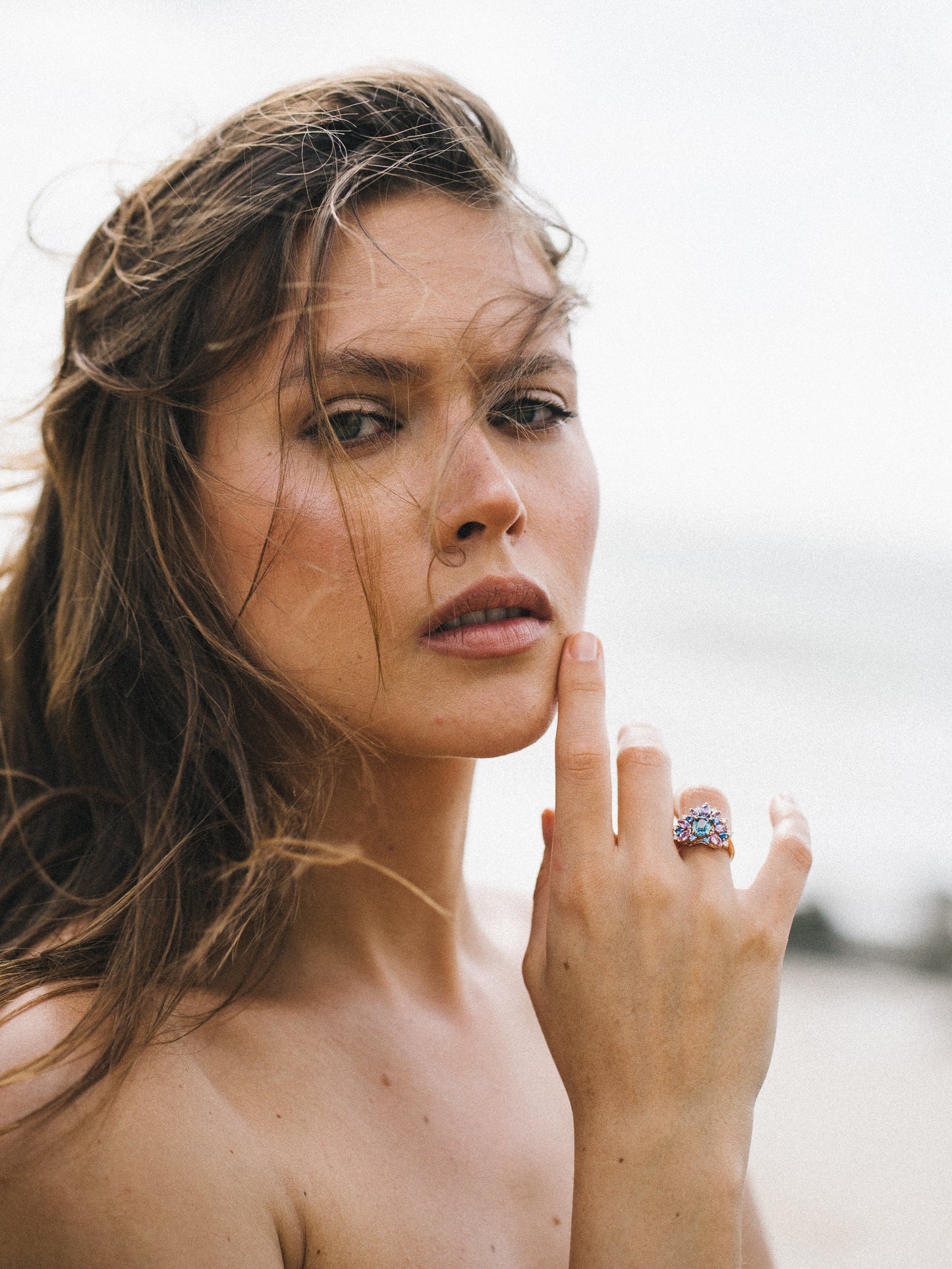 Une personne aux longs cheveux bruns se tient dehors, regardant droit vers l'objectif. Elle se touche le menton, mettant en valeur une bague colorée Indigo Bloom - Saphir de Bonnot Paris. L'arrière-plan présente une scène naturelle floue.