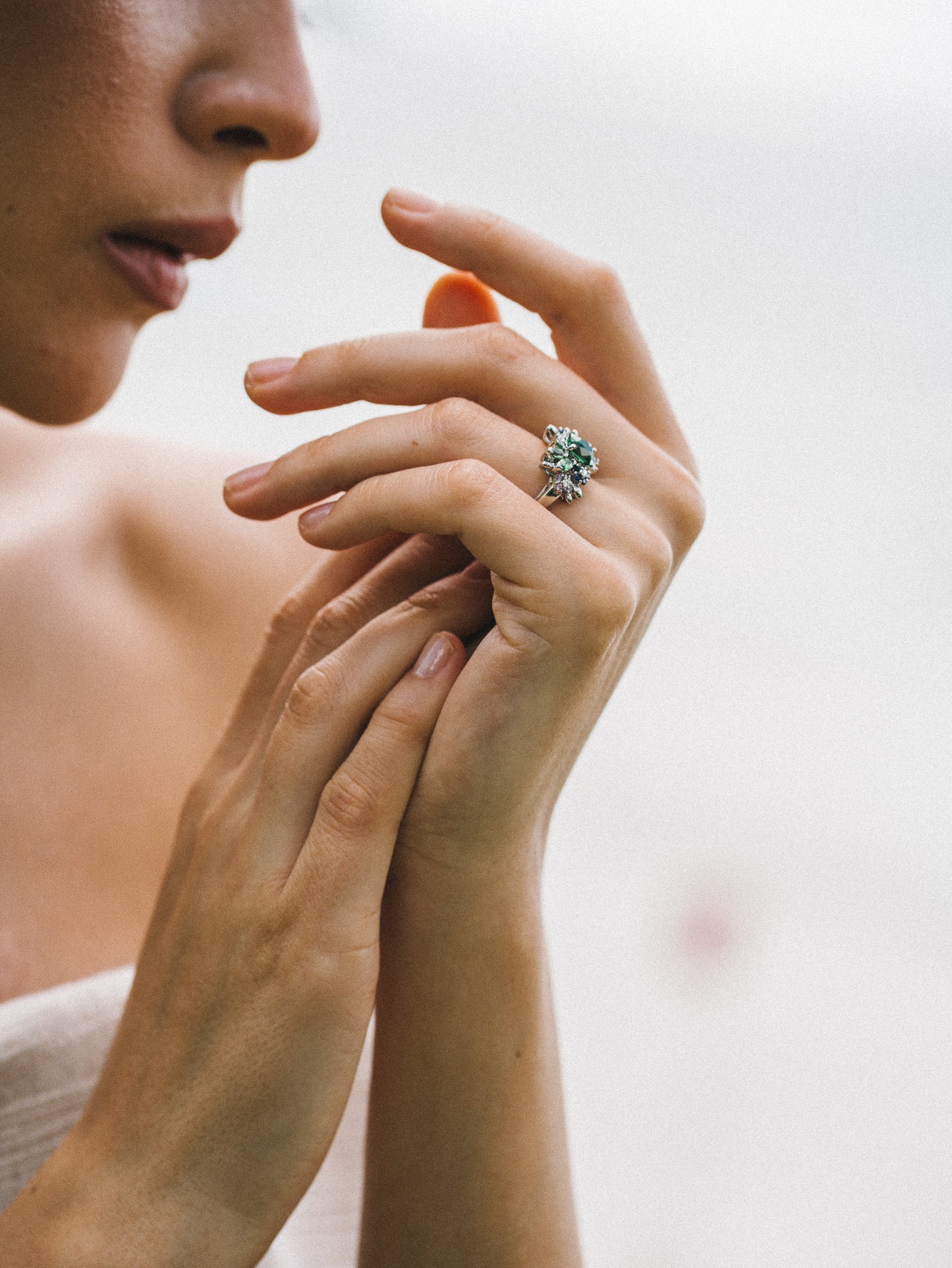 Une femme touche délicatement ses mains, mettant en valeur la collection Indigo Bloom - Saphir de Bonnot Paris, ornée d'une large bague florale sertie de diamants scintillants à l'annulaire droit. Le fond doux souligne sa grâce et la raffinement de sa haute joaillerie.