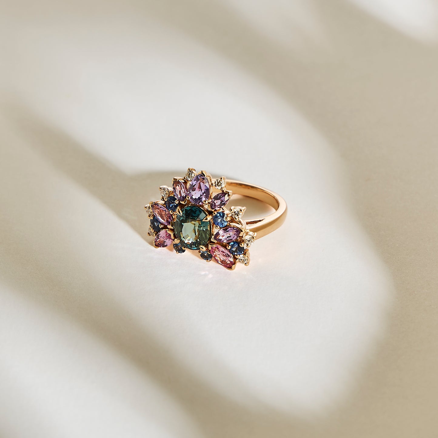 La bague Indigo Bloom - Saphir de Bonnot Paris présente une surface en or lisse ornée d'un amas de pierres violettes, vertes et roses. Les ombres créent un effet texturé, incarnant l'élégance et le luxe de la haute joaillerie.
