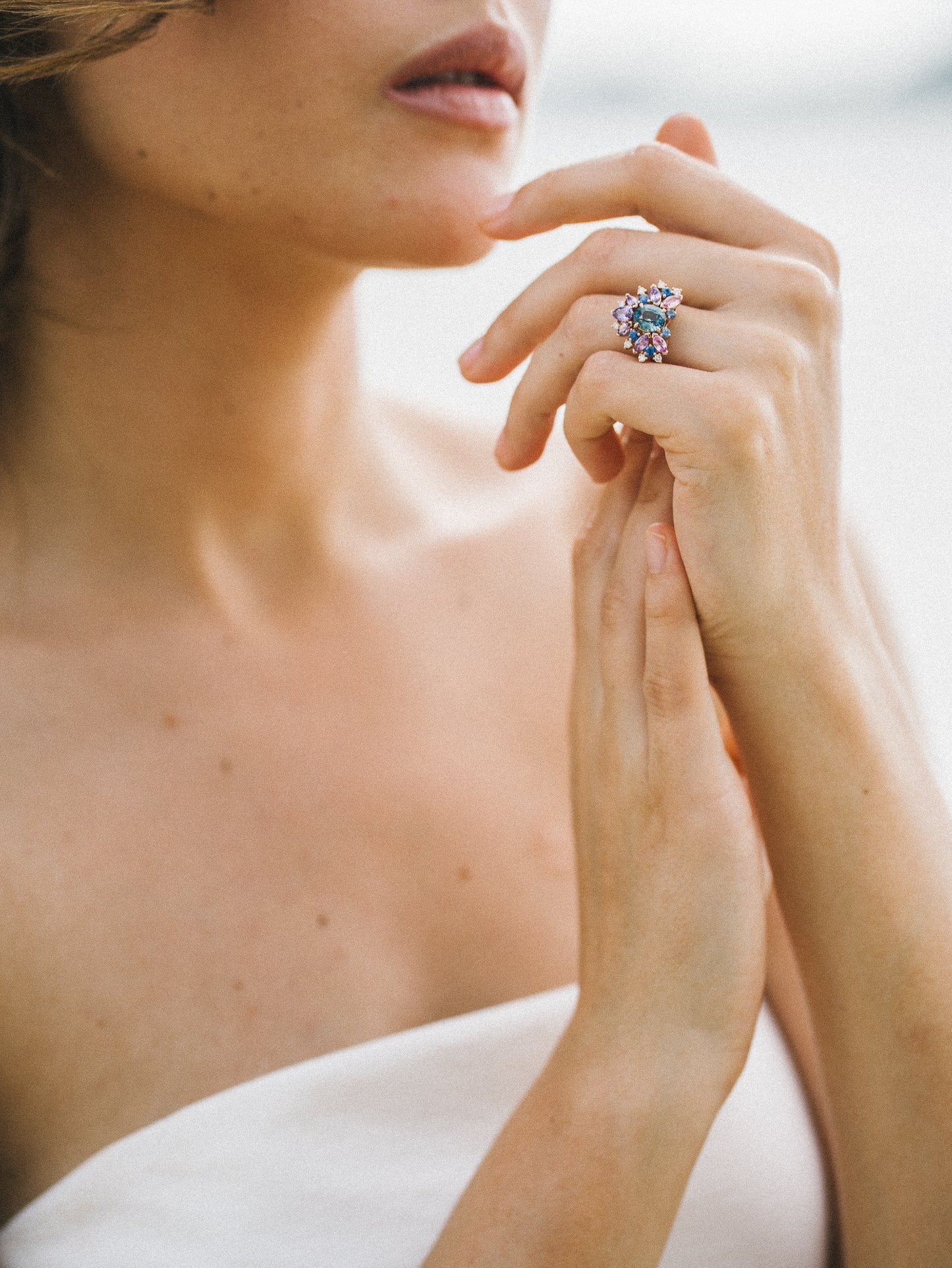 Une femme aux épaules nues pose avec élégance, les mains près du visage, exhibant la bague Indigo Bloom - Saphir de Bonnot Paris. Les pierres précieuses colorées et le saphir bleu haute joaillerie lui confèrent une touche de luxe et d'élégance.