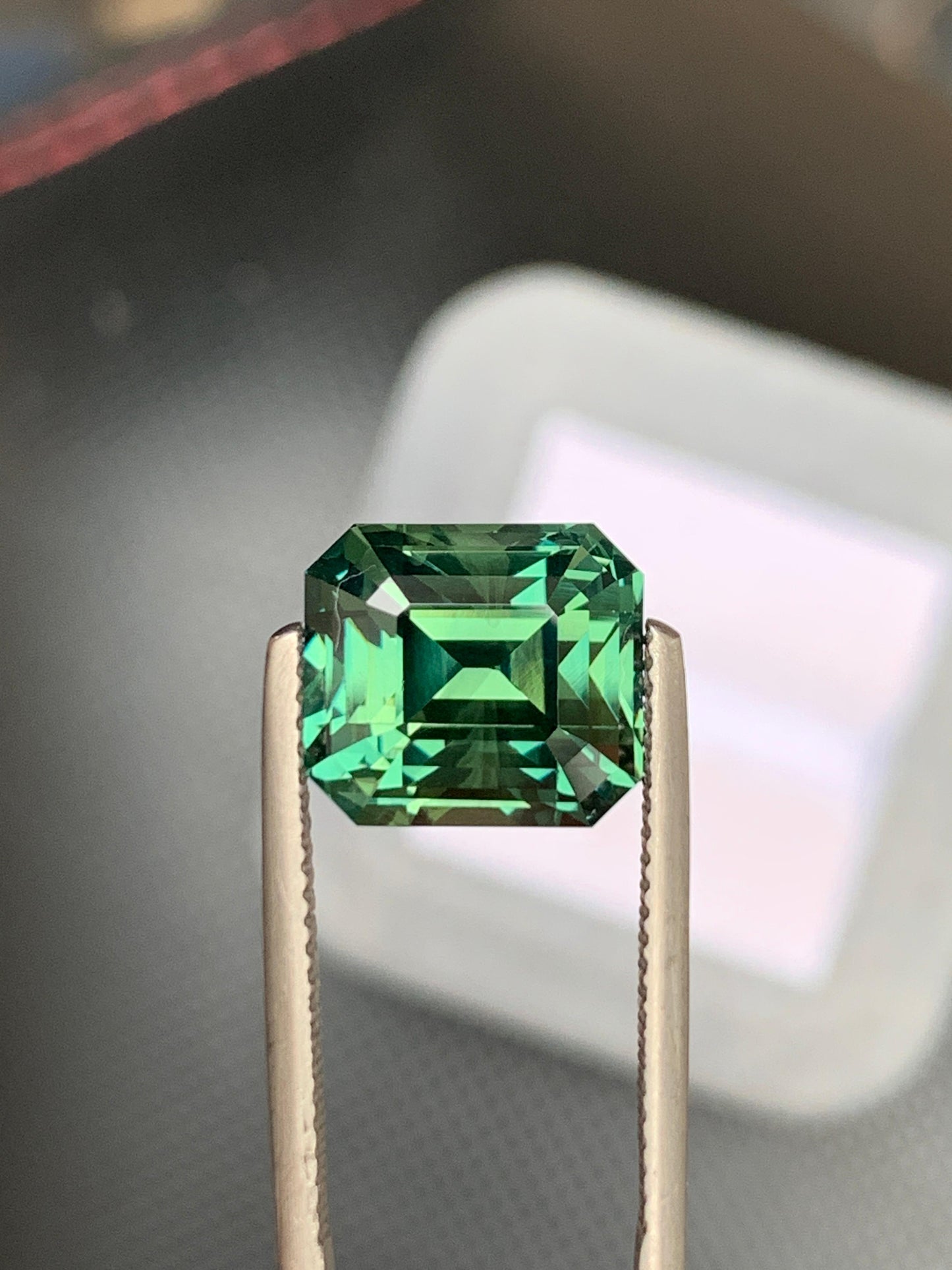 Une pince à épiler tient le Saphir vert • 10,55 carats de Bonnot Paris, une pierre précieuse taille émeraude éclatante de Madagascar. Cette pierre rectangulaire de 10,55 carats présente des facettes étagées qui mettent en valeur sa teinte verte profonde et brillante sur un fond flou.