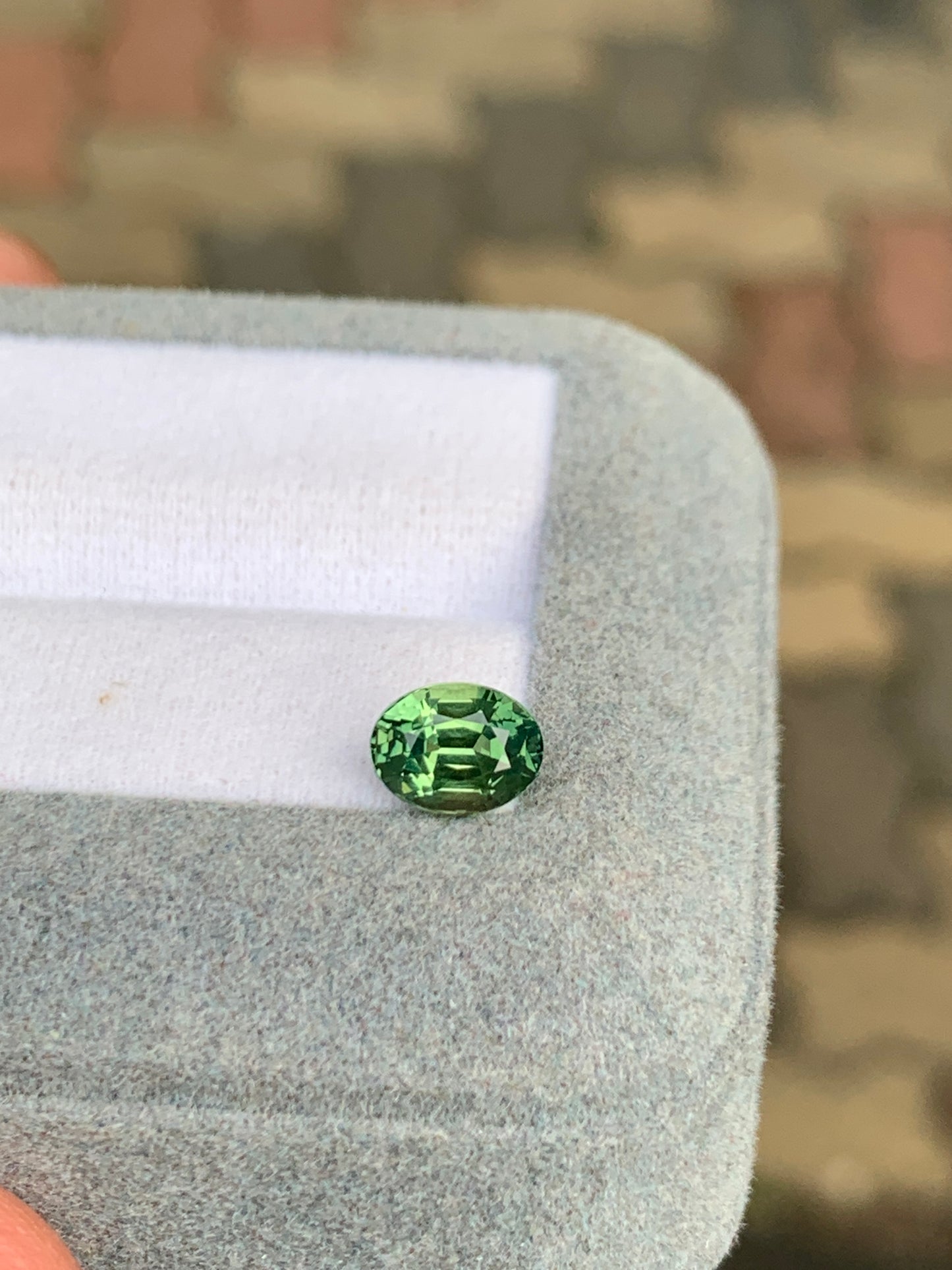 Gros plan sur le Saphir vert de Madagascar de 1,55 ct de Bonnot Paris, posé dans un coffret bleu-gris clair avec un insert en tissu blanc. Un motif flou en arrière-plan rehausse l'attrait de la pierre précieuse.