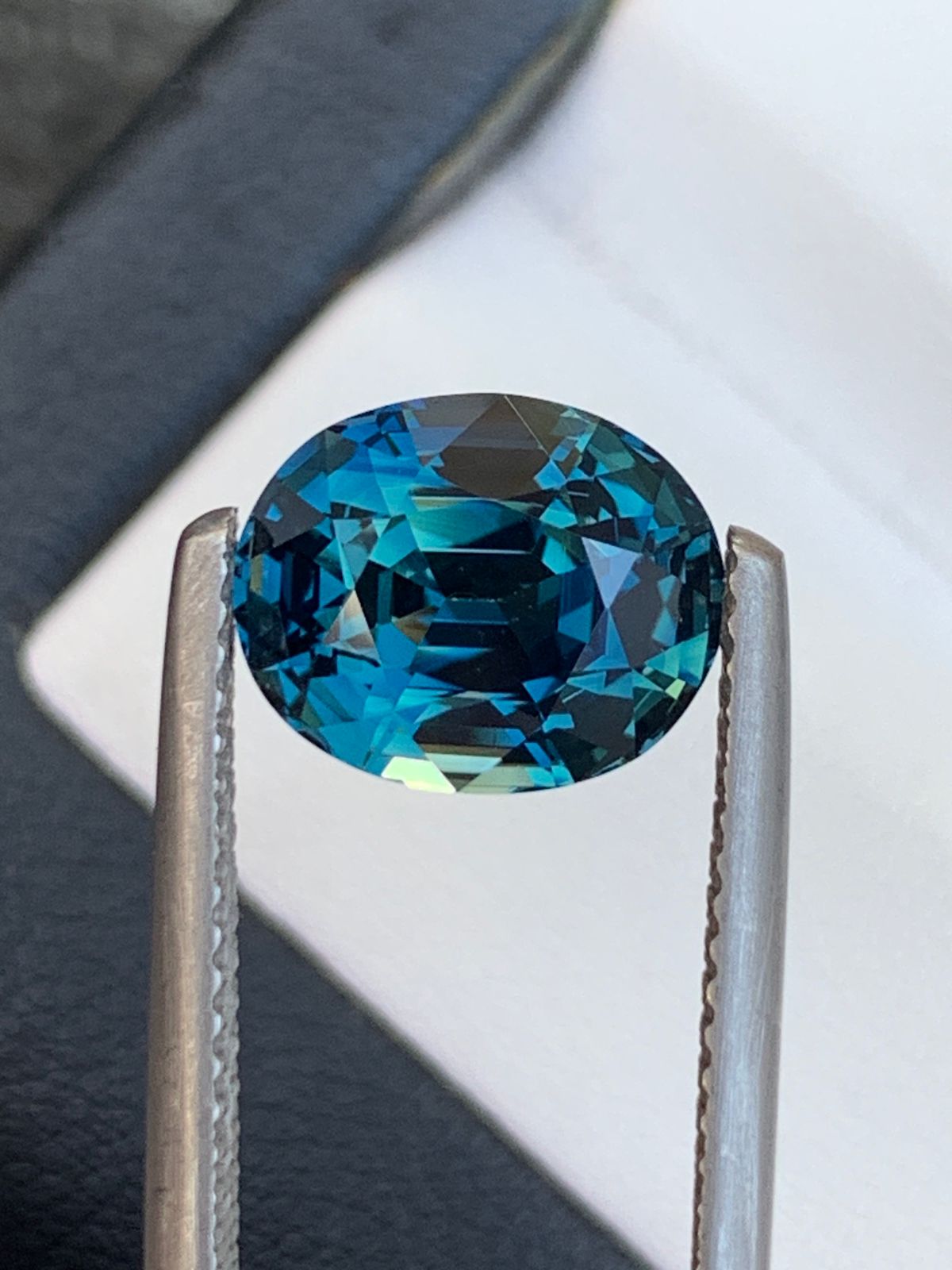 Gros plan de la pierre précieuse Saphir Teal de Bonnot Paris de 4,1 carats, photographiée à la pince à épiler sur un fond flou. Cette pierre ovale présente des facettes complexes qui reflètent des nuances fascinantes de bleu et de vert, idéales pour une bague de fiançailles saisissante.