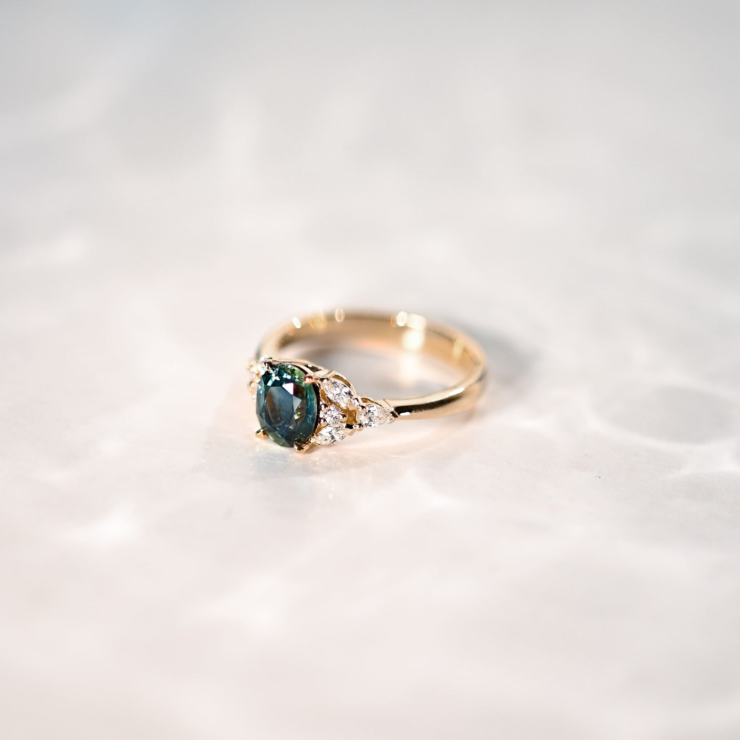 La Bague Saphir Teal · 1,5 carat de Bonnot Paris repose sur une surface en marbre. Sa pierre centrale turquoise, rehaussée de petites pierres transparentes, reflète une douce lumière, soulignant son charme floral subtil.