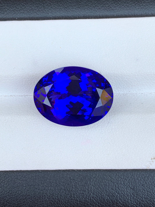 Une tanzanite bleue facettée de 21,3 carats, de forme ovale et éclatante, signée Bonnot Paris, est mise en valeur sur une surface blanche. Cette pierre précieuse présente des teintes bleu profond avec des reflets violets et complexes, élégamment positionnées sur un fond noir et blanc saisissant.