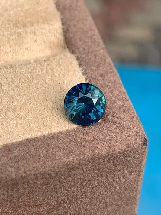 Un saphir sarcelle rond de 2,4 carats de Madagascar de Bonnot Paris repose sur une surface brune texturée, mettant en valeur son bleu profond avec des reflets verts sur un fond bleu flou.