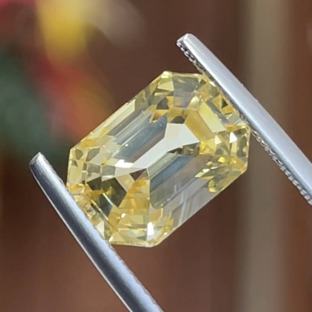 Un gros plan du Saphir Jaune · 7,95ct de Bonnot Paris montre ses facettes taille émeraude jaune claire et sa transparence, maintenues par une pince à épiler sur un fond flou.