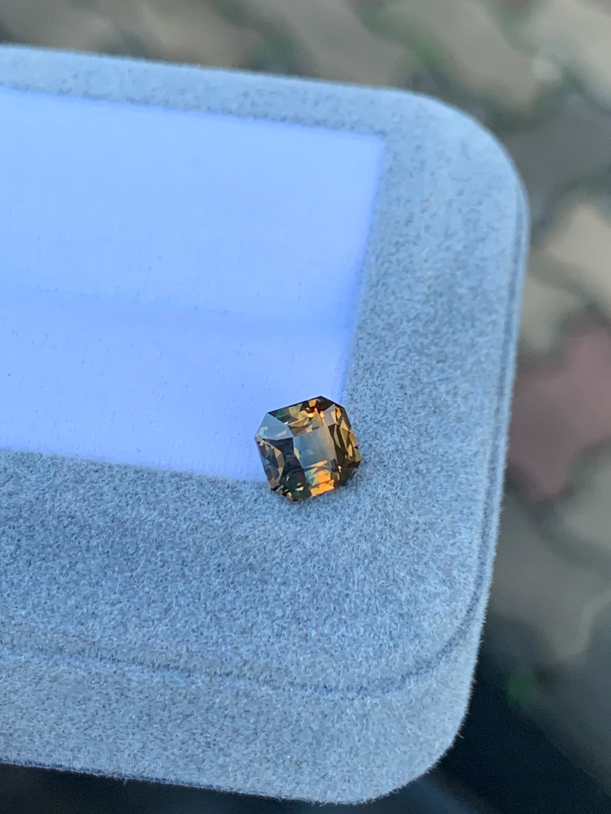 Un Saphir bicolore de 1,60 ct de [NOUVEAU] Bonnot Paris, aux tons ambrés et multifacettes, repose avec une lueur orange chaleureuse sur le coin d'une boîte à bijoux grise à la texture douce sur un fond flou.
