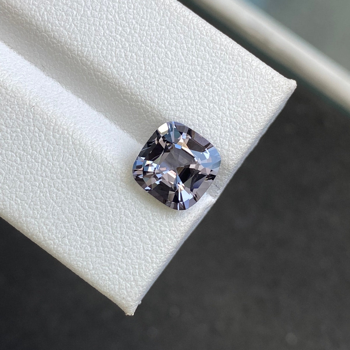La pierre précieuse Spinelle Gris 3,31 ct de Bonnot Paris, avec sa taille coussin et sa teinte gris-violet, est présentée sur une surface blanche texturée, capturant la lumière pour révéler ses facettes et ses reflets sur un fond gris foncé légèrement flou.