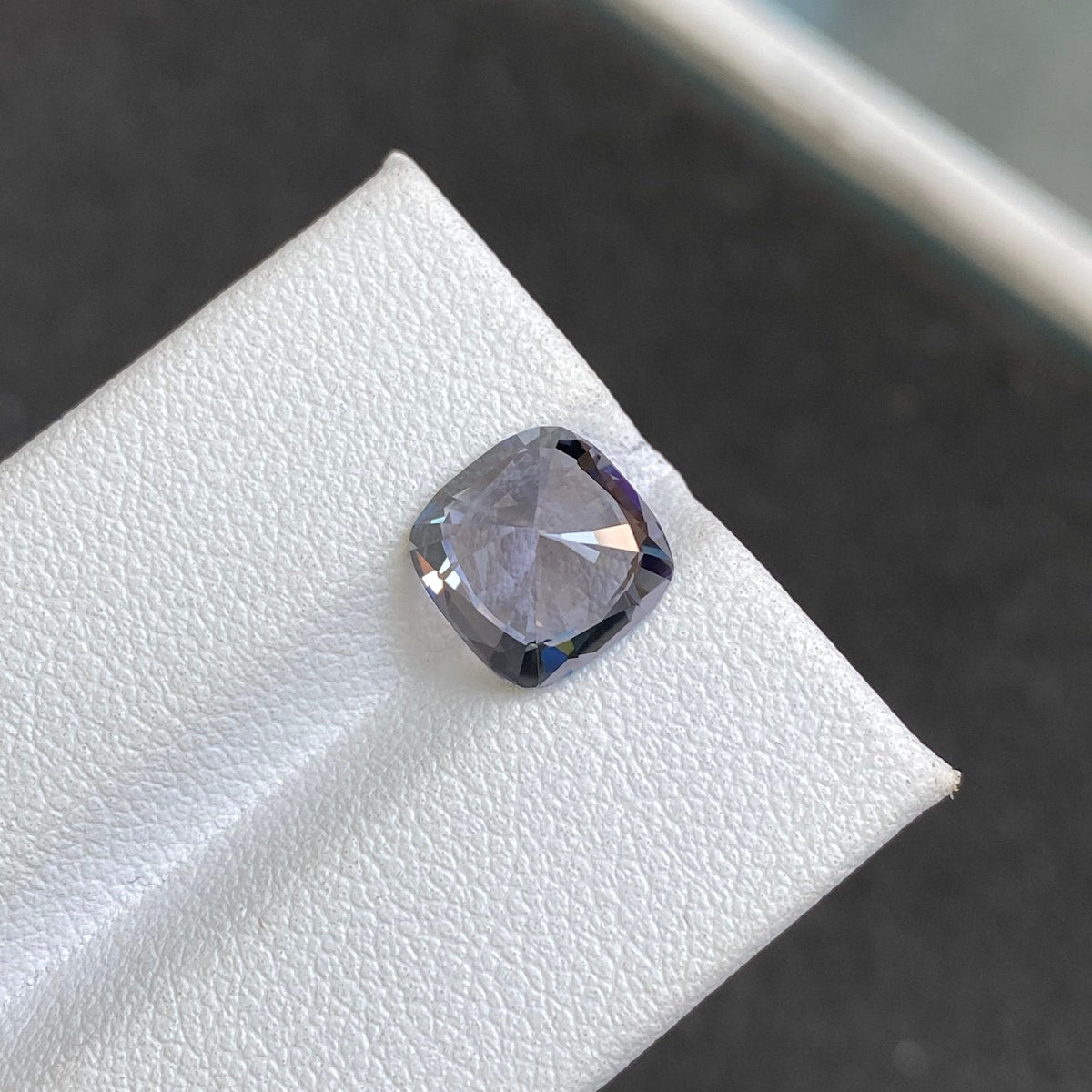 Le Spinelle Gris Bonnot Paris, une pierre transparente taille coussin de 3,31 carats, se présente sur une surface blanche sur fond sombre. Cette pierre facettée reflète élégamment la lumière avec un éclat subtil.