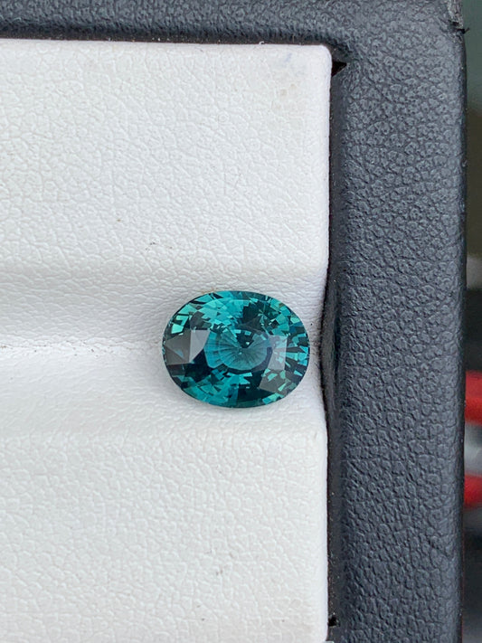 Gros plan d'une pierre précieuse ronde facettée Saphir Teal • 3,05 ct de Bonnot Paris sertie sur un fond blanc et gris, mettant en valeur ses teintes vibrantes bleu sarcelle et gris foncé alors qu'elle reflète la lumière.
