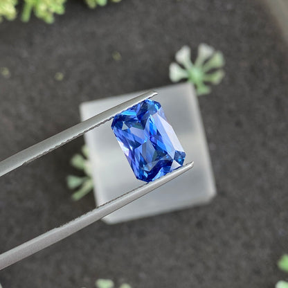 Une paire de pinces maintient la pierre précieuse vibrante et facettée Saphir Bleu · 4,13 ct de Bonnot Paris sur un fond flou avec de petites plantes vertes, soulignant sa clarté et sa couleur riche.