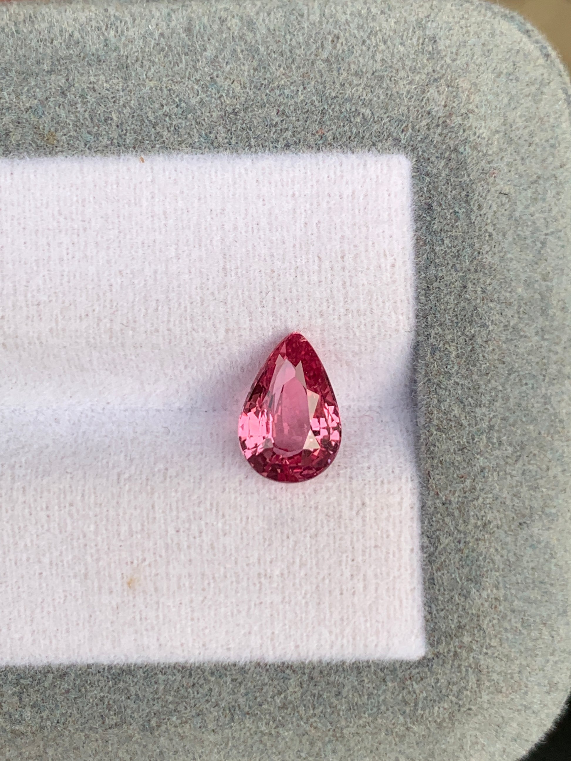 Une gemme Saphir Padparadcha en forme de poire (2,04 ct) de Bonnot Paris, avec sa surface facettée scintillante, présentée sur un tissu blanc doux à l'intérieur d'une boîte à bijoux gris-vert.