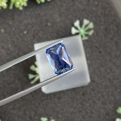 Une paire de pinces en métal maintient le Bonnot Paris Saphir Bleu · 4,13 ct, une pierre précieuse bleue taille émeraude, sertie sur un fond flou et sombre avec des touches de verdure pour un contraste naturel.
