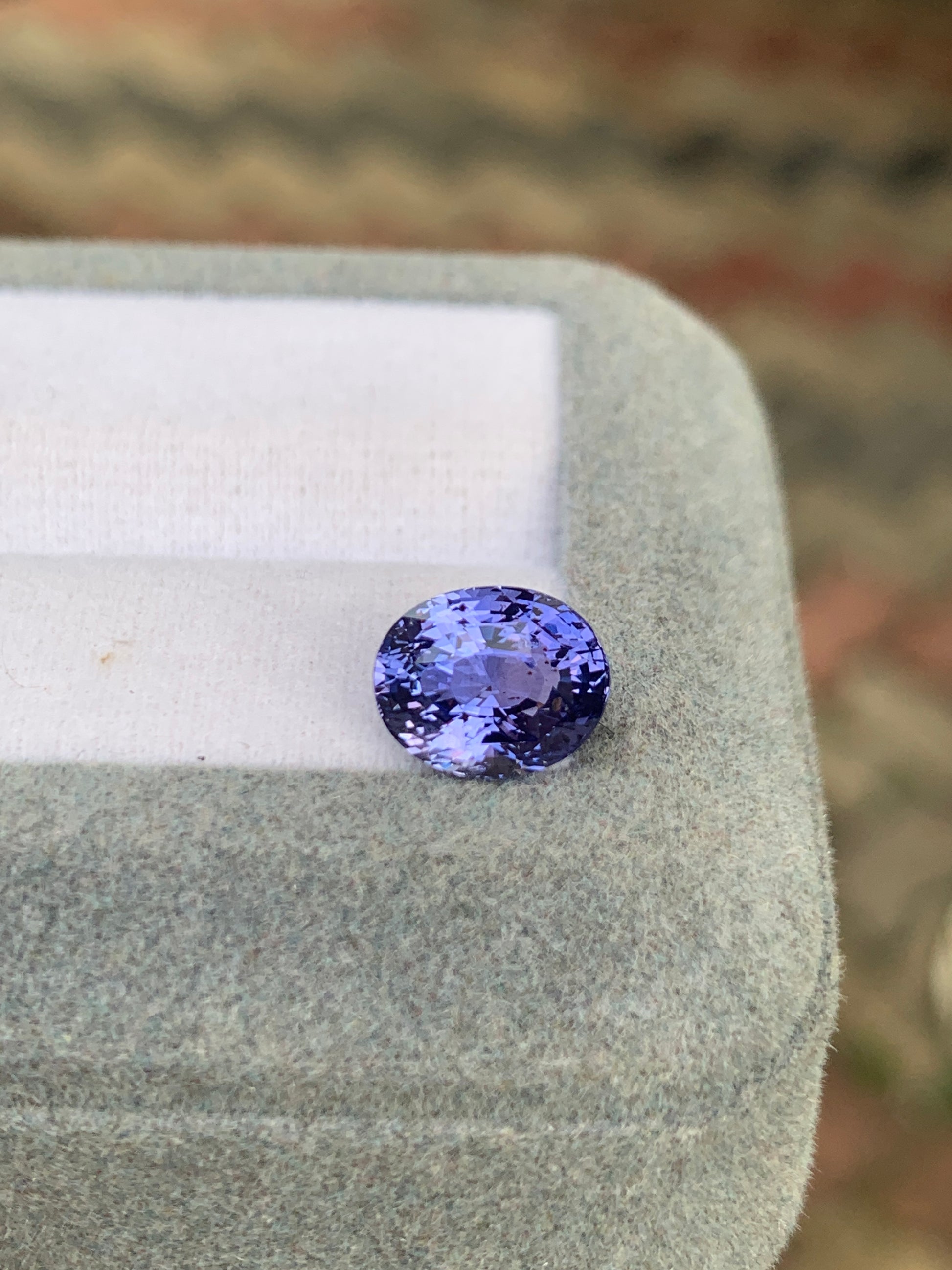 Une pierre précieuse saphir violet de 3,52 carats de Bonnot Paris, originaire du Sri Lanka, repose sur un doux coussin gris clair. Ses teintes lavande et ses facettes captivantes se détachent sur un fond flou aux tons terreux, accentuant son charme discret.