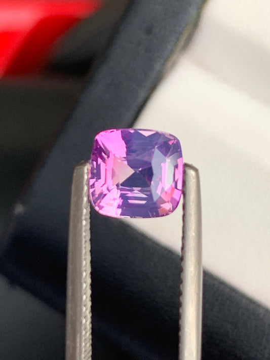 Un gros plan du Saphir Violet • 2,08ct de Bonnot Paris, une pierre précieuse rose facettée rectangulaire maintenue par une pince en argent, évoque une délicate pierre précieuse du Sri Lanka sur un fond flou de teintes sombres et claires.