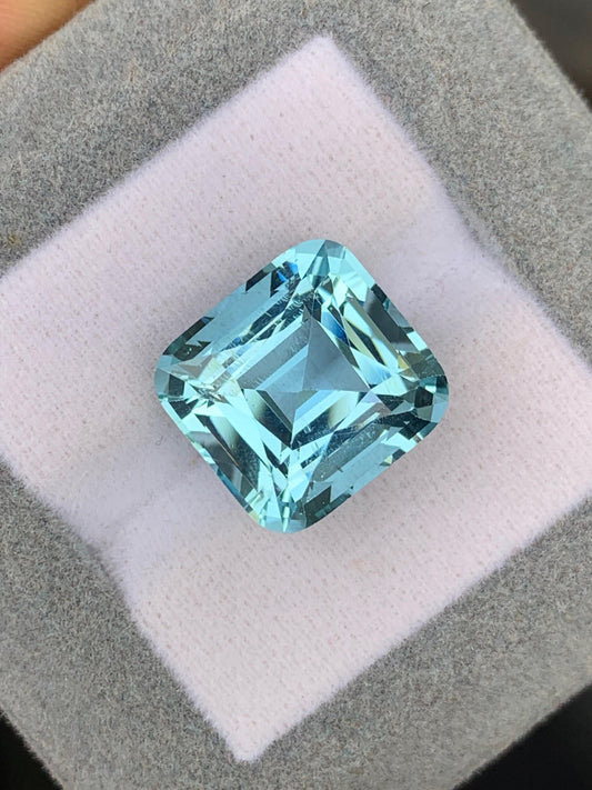 Un gros plan sur l'Aigue-Marine • 12,58ct deBonnot Paris révèle ce bijou unique aux facettes reflétant la lumière dans des nuances de bleu envoûtantes, sur fond gris et blanc coussiné.
