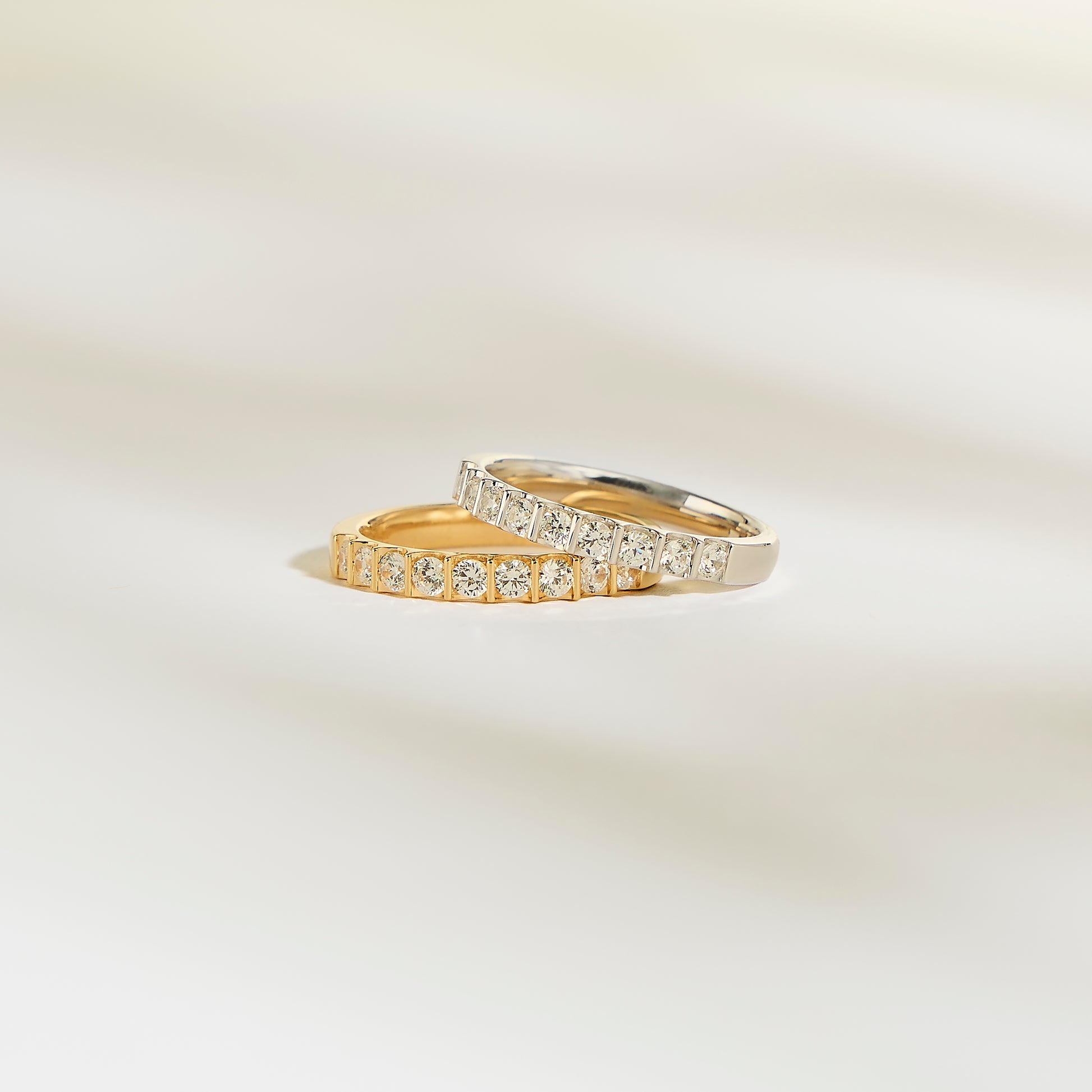Les bagues Alliance La Majestueuse de Bonnot Paris, l'une en or et l'autre en argent, arborent des diamants brillants sur une surface douce et claire. Le fond légèrement flouté rehausse le luxe serein de la couleur clarté G/H SI.