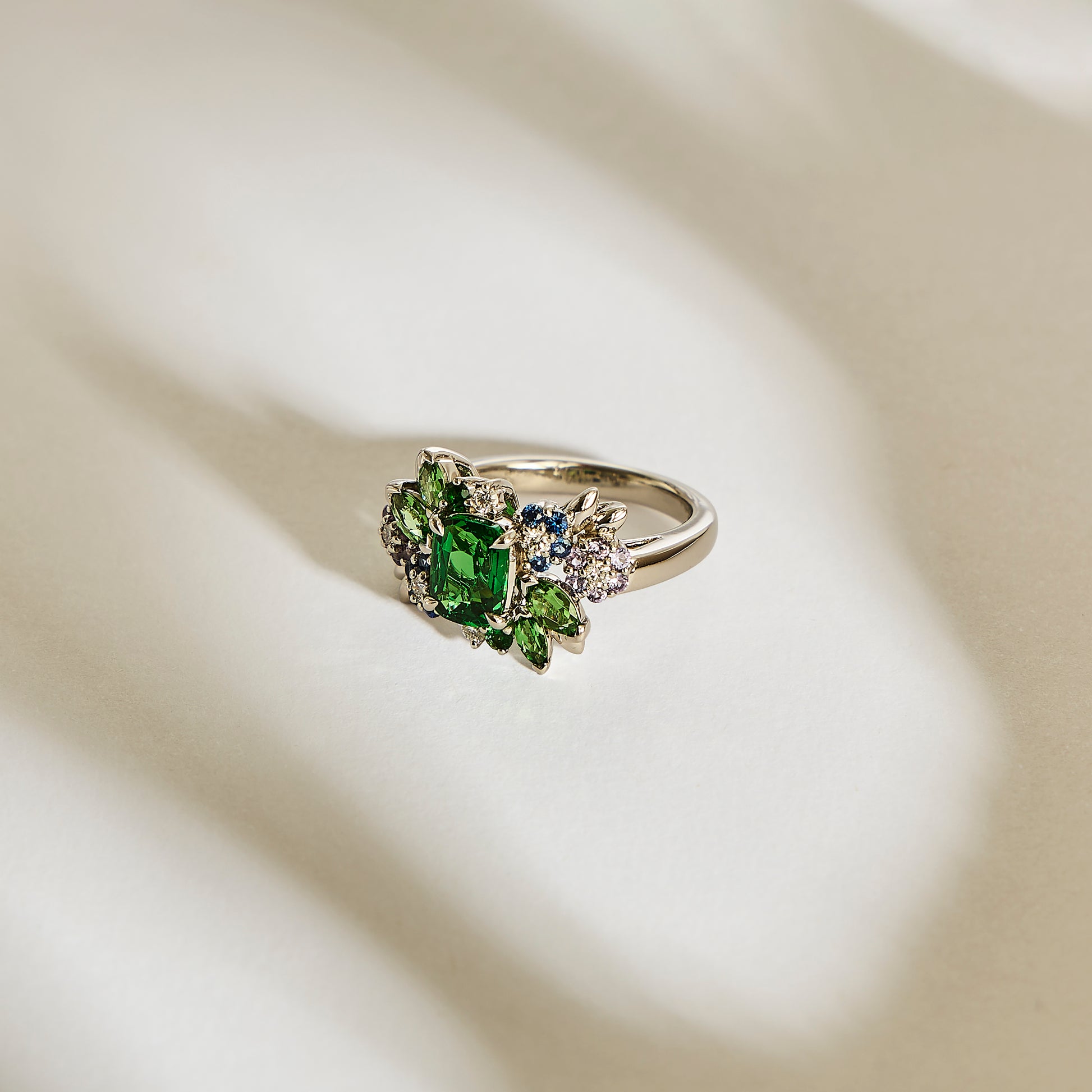 La bague en or Green Bloom - Tsavorite de Bonnot Paris présente une grande pierre précieuse verte entourée de plus petites tsavorites Grenat et de pierres claires, élégamment présentées sur une surface légèrement texturée et de couleur claire avec des ombres douces.