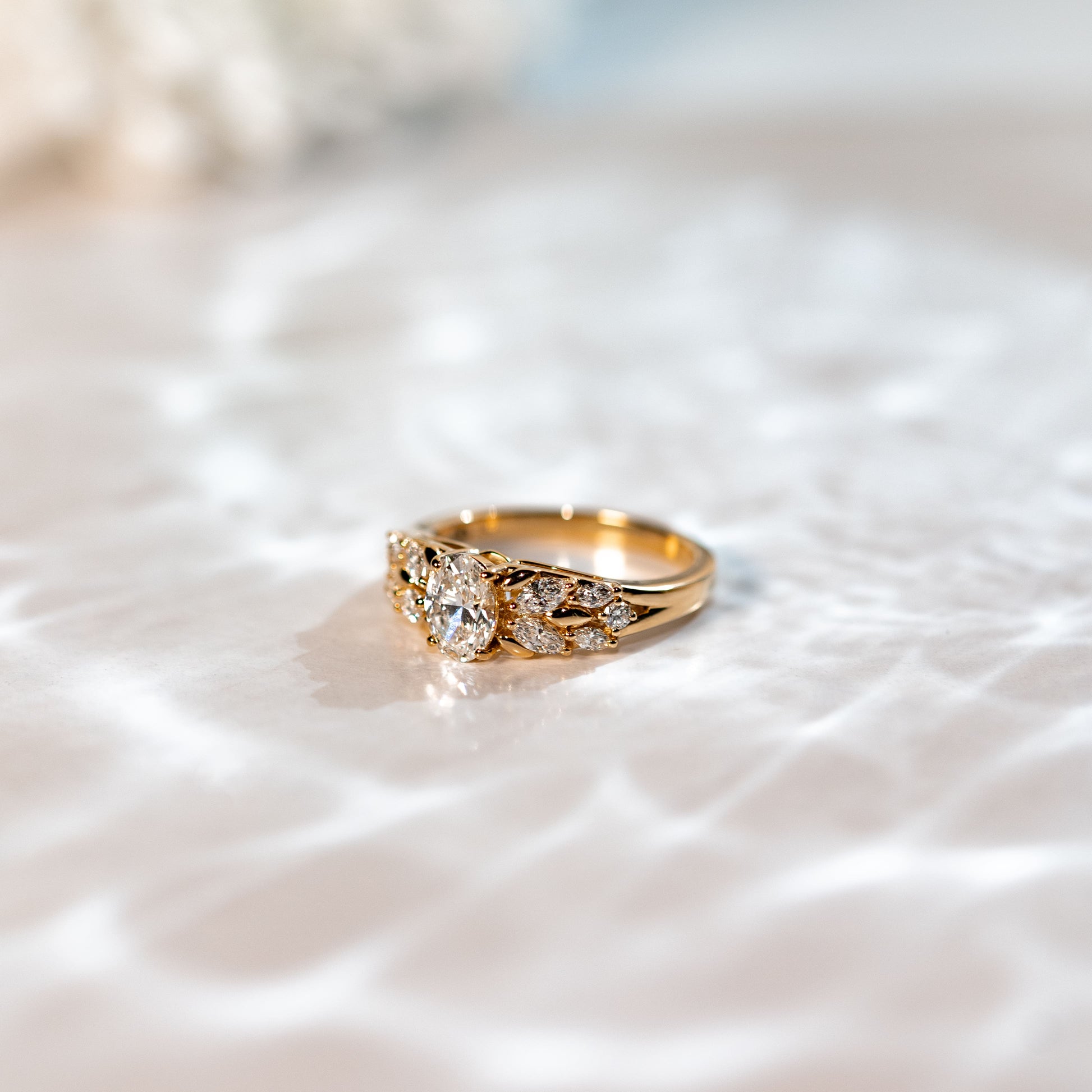 La Bague Diamants · 0,7 carat de [NOUVEAU] Bonnot Paris, ornée d'un diamant ovale central et de diamants plus petits, repose sur une surface blanche texturée. La lumière naturelle rehausse la sophistication de la bague par de doux reflets.