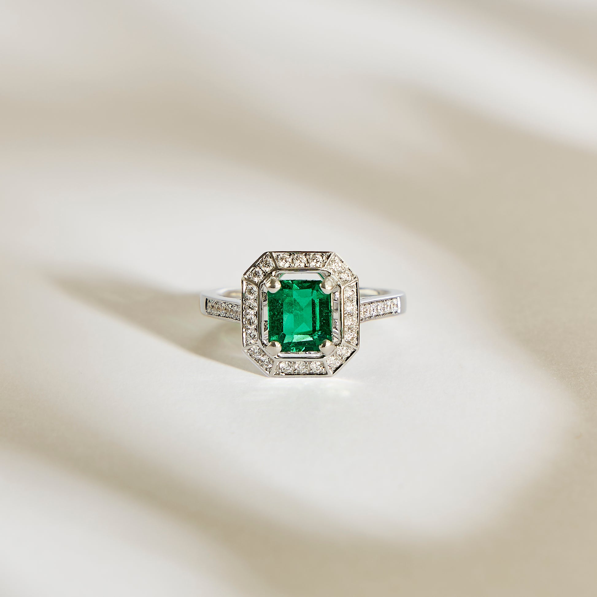 La Bague Emeraude • 1,22ct de Bonnot Paris présente une pierre précieuse verte taille émeraude évoquant le style Art Déco, entourée d'un halo de diamants étincelants sur une surface argentée aux ombres douces.
