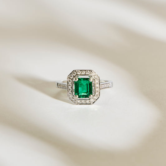 La Bague Emeraude • 1,22ct de Bonnot Paris présente une pierre précieuse verte taille émeraude évoquant le style Art Déco, entourée d'un halo de diamants étincelants sur une surface argentée aux ombres douces.