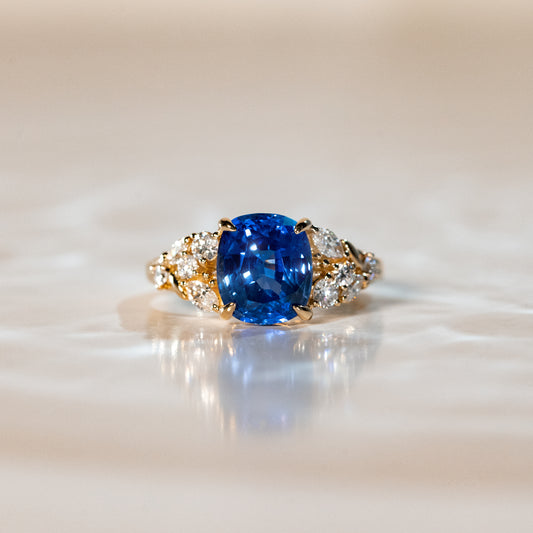 La Bague Saphir • 3,51 carats de [NOUVEAU] Bonnot Paris arbore un design en or orné d'une pierre précieuse ovale bleue rappelant les teintes saphir et bleuet, flanquée de grappes de diamants. Elle repose élégamment sur une surface réfléchissante pâle, sur un fond légèrement flou.