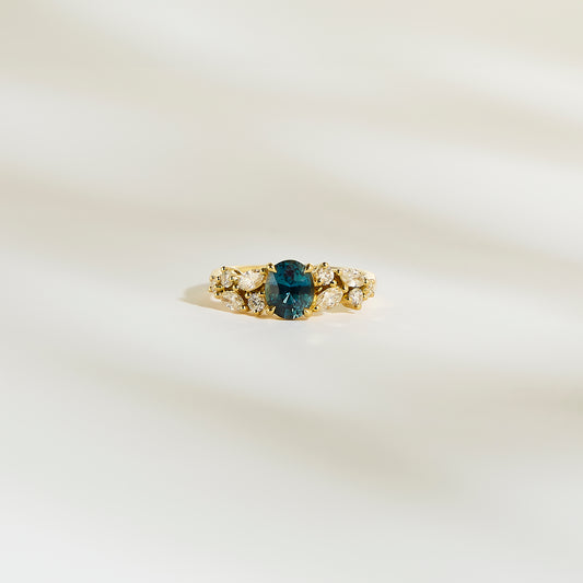 Sur une surface douce et lisse de couleur crème se trouve la « Bague Saphir Teal • 1,14ct » de Bonnot Paris, comportant un saphir bleu-vert ovale en son centre entouré de petits diamants blancs étincelants.
