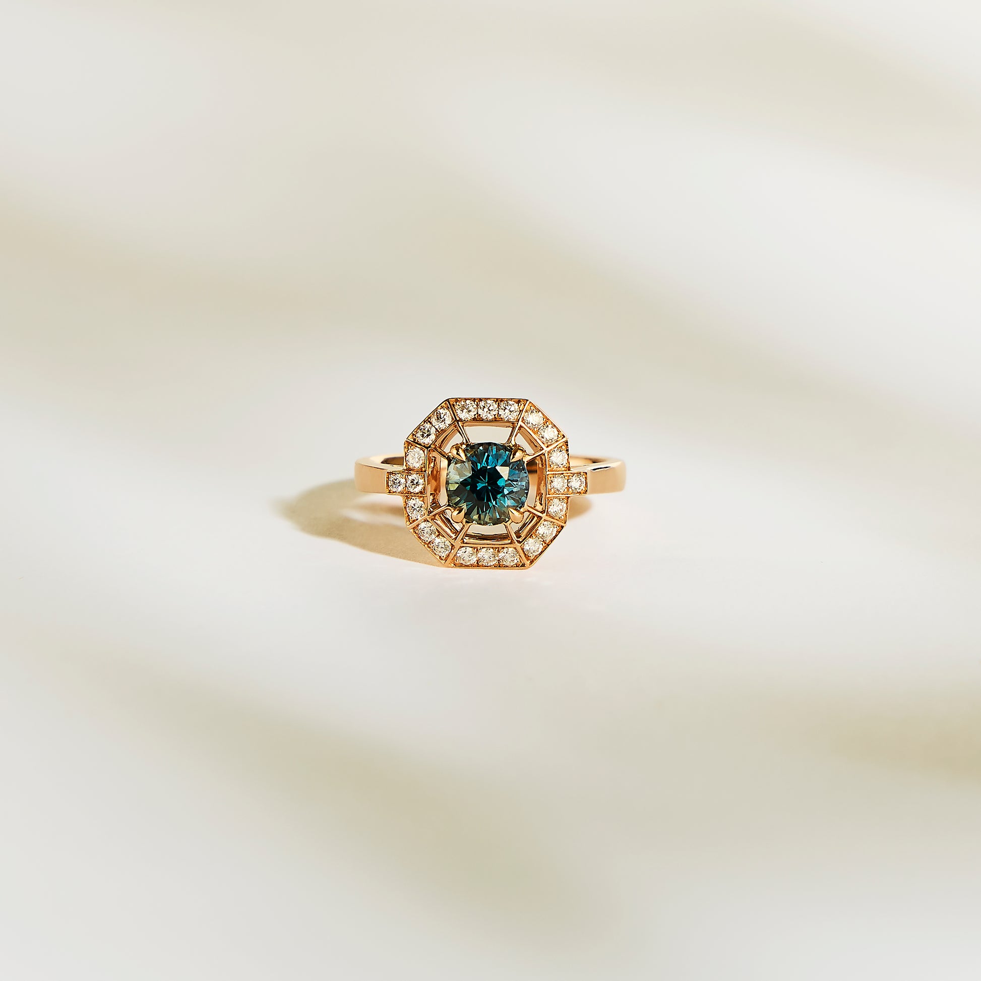 La Bague Saphir Teal de Bonnot Paris • 1,16 ct est une superbe bague de fiançailles en or avec un saphir sarcelle central et un halo de diamants blancs étincelants, présentés sur une surface douce et claire.