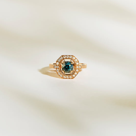 La Bague Saphir Teal de Bonnot Paris • 1,16 ct est une superbe bague de fiançailles en or avec un saphir sarcelle central et un halo de diamants blancs étincelants, présentés sur une surface douce et claire.