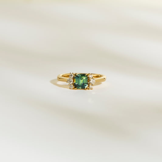 La Bague Bonnot Paris Saphir Teal • 1,24 ct présente une pierre précieuse sarcelle carrée flanquée de petits diamants blancs étincelants sur une bande lisse et de couleur claire.