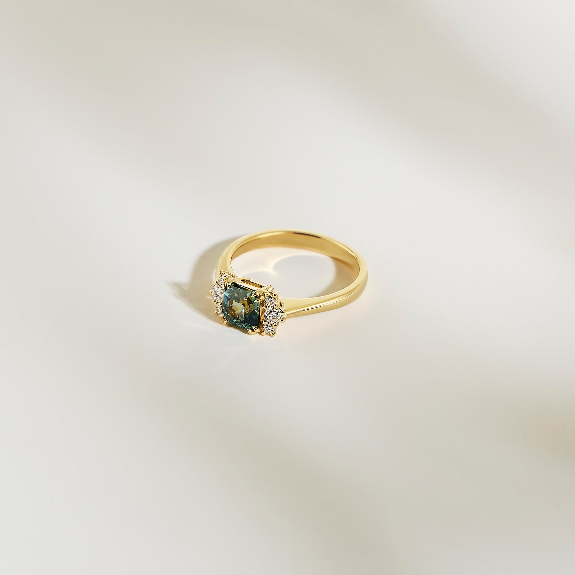 La Bague Saphir Teal • 1,24ct de Bonnot Paris, une bague en or avec un saphir sarcelle taille carrée en son centre et entouré de petits diamants blancs, est présentée sur une surface lisse de couleur claire.