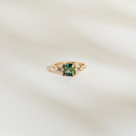 La Bague Saphir Teal • 1,56 ct de Bonnot Paris est une bague de fiançailles en or présentant un saphir bleu-vert rectangulaire, flanqué de petits diamants blancs sur un fond blanc doux.